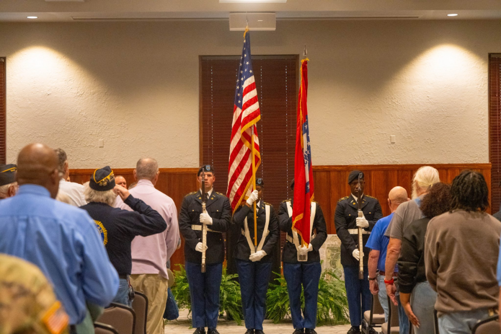 Army ROTC Color Guard stands at attention in front of a podium. Audience members on either side of them stand tall, veterans salute the flag.