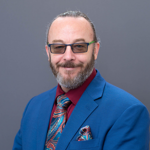 Professional headshot of Wade Maki, a man with light skin, glasses, and a neatly groomed beard and mustache. He is smiling slightly and wearing a bright blue suit jacket, a burgundy shirt, and a colorful paisley tie with a matching pocket square. His hair is pulled back, and the photo is set against a neutral gray background.