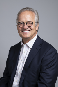 A professional headshot of a smiling middle-aged man with gray hair and glasses, wearing a navy blazer and a light-colored button-down shirt, against a neutral gray background.
